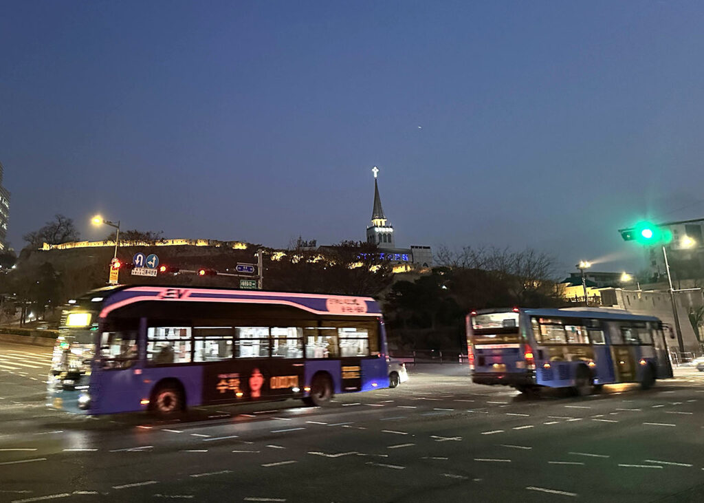 Imagem mostra fragmento da antiga muralha de Seul ao fundo e dois ônibus em destaque.